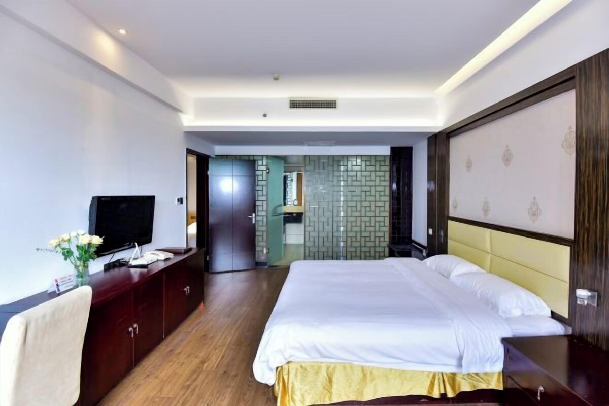 A hotel room with a large bed, a television, a desk with a vase, and a wardrobe.