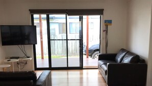 A living room with a black leather sofa, a wooden dining table, and a TV on a stand. - Seabreeze Escape | Pet-Friendly Coastal Retreat | Sleeps 8 | Walk to Beach (Cape Paterson)
