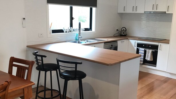 A modern kitchen with a central island, bar stools, a dining table, and a window with a 'welcome' sign. - Seabreeze Escape | Pet-Friendly Coastal Retreat | Sleeps 8 | Walk to Beach (Cape Paterson)
