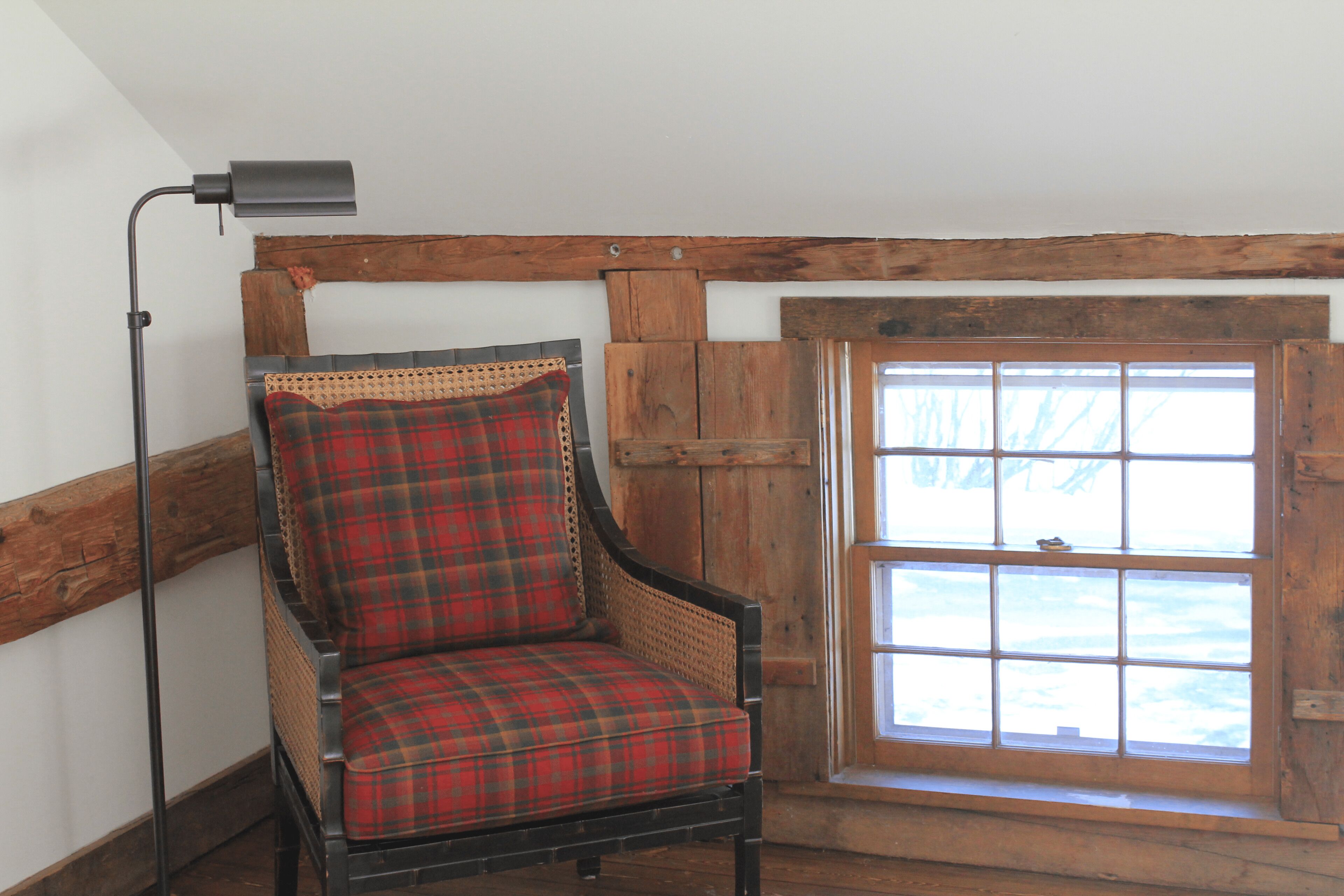 A corner room with a plaid chair, a floor lamp, and a wooden window with a view.