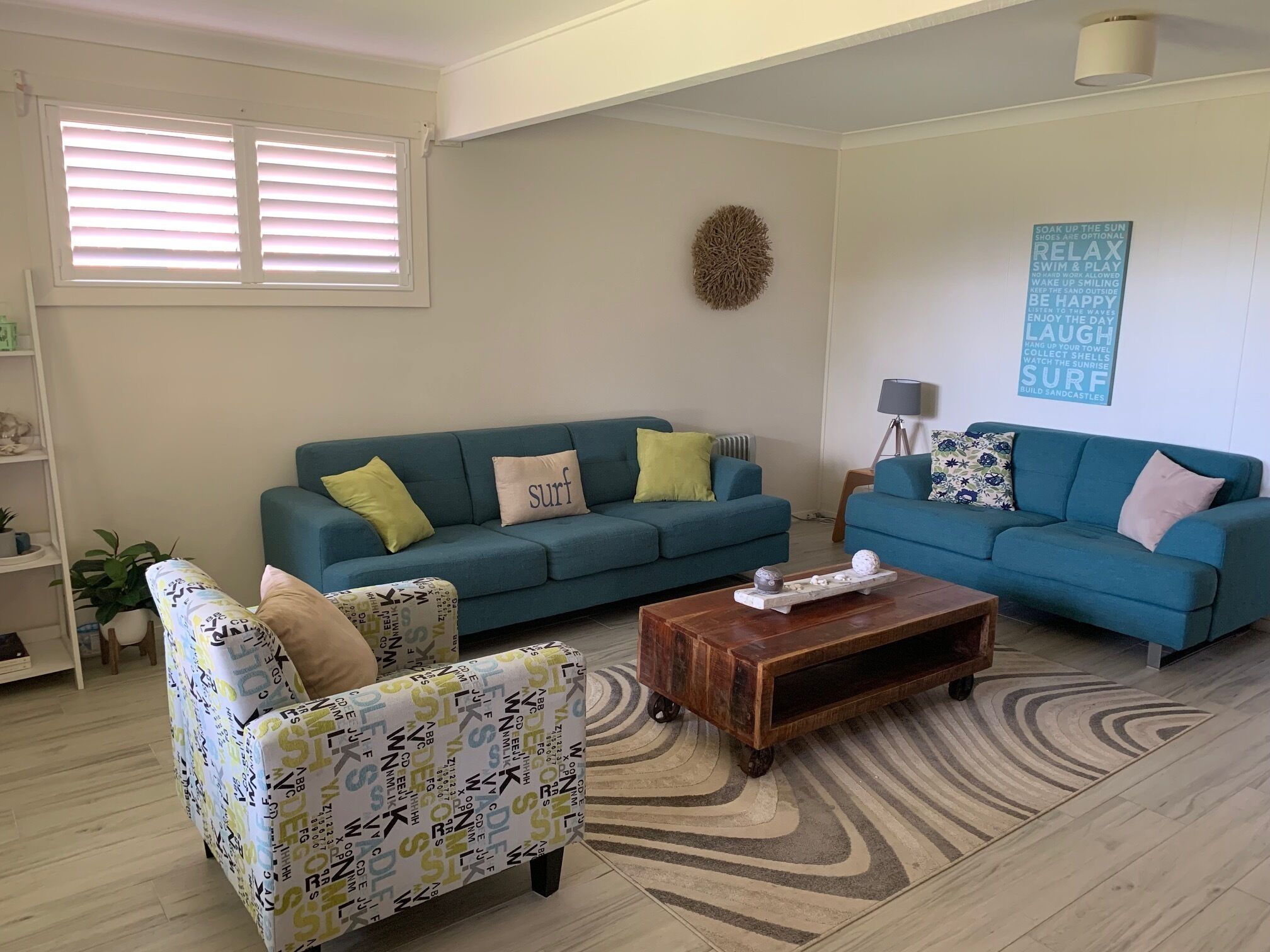 Una sala de estar con dos sofás azul verdosos, un sillón estampado, una mesa de centro de madera y un adorno en la pared.