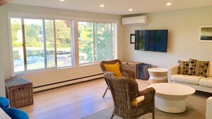 A spacious living room with large windows, wicker furniture, a white sofa, and a flat-screen TV. - Central Woods Hole Village Waterfront! (Falmouth)