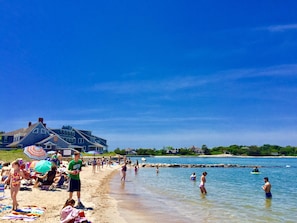 A beach with people, umbrellas, and a large house in the background.