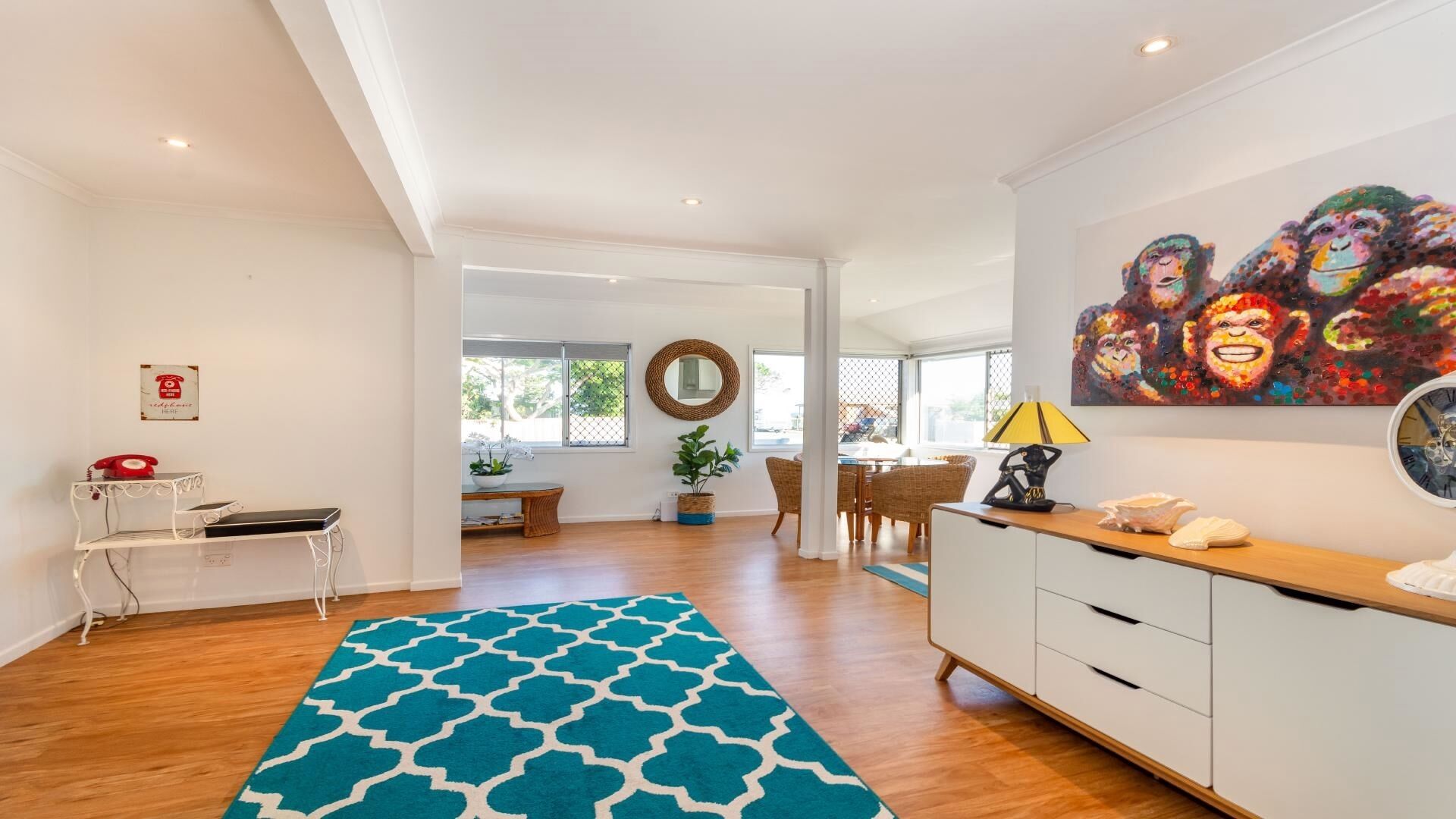A spacious living area with a large blue patterned rug, a dining table with chairs, a white cabinet, and a colorful painting on the wall.