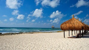Une plage avec des parasols en chaume et des chaises longues, une jetée en arrière-plan et un ciel bleu dégagé.