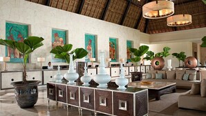 Un hall d’entrée spacieux avec un plafond haut, un comptoir de réception, de grandes plantes en pot et une table basse centrale.