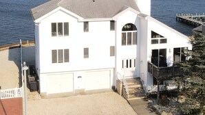 A white two-story house with a garage, a covered porch, and a view of the water and a bridge in the background. - Bayfront Home with Pier, Multi-Level Decks & Stunning LBI Views (Manahawkin)
