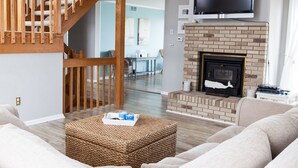 A living room with a fireplace, a television mounted above it, and a staircase leading to another room. - Bayfront Home with Pier, Multi-Level Decks & Stunning LBI Views (Manahawkin)