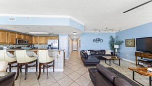 A modern kitchen with a central island, a dining area with bar stools, a living room with a leather sofa and a TV, and a hallway leading to a bedroom.