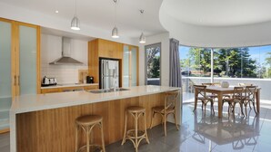 A modern kitchen with a central island, wooden cabinets, stainless steel appliances, and a dining area with wicker chairs. - The White House - Merimbula (Merimbula)