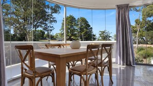 A dining area with a wooden table and chairs, large windows with a view of trees and a car outside. - The White House - Merimbula (Merimbula)