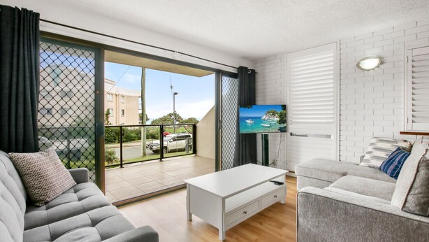 Una sala de estar moderna con un sofá gris, una mesa de centro blanca y un sillón gris. Un televisor está montado en la pared, y se observa un balcón con vistas a la calle y edificios.