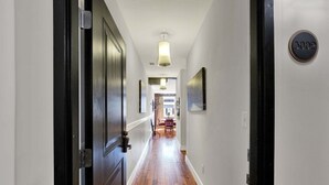 Un pasillo con suelo de madera, una puerta con placa de número y una lámpara colgante.