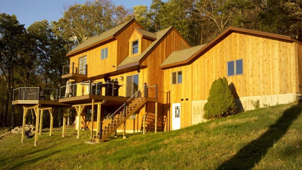 A two-story wooden cabin with a balcony on the upper level, a covered outdoor seating area, and a staircase leading to the upper floor. - Magnificent Riverfront home, completely private.  BOOK  7 NIGHTS GET 10% OFF (Middle Haddam)