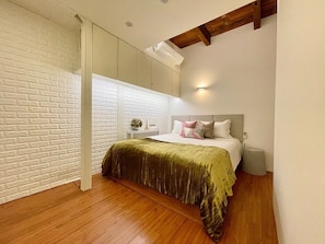 Un dormitorio con piso de madera, una cama con colcha verde, pared de ladrillo blanco y viga de techo de madera.