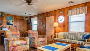 A cozy living room with wooden walls, a striped sofa, colorful armchairs, a wooden coffee table, and a ceiling fan.