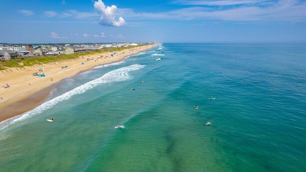An aerial view of a beach with clear water, a sandy shore, and buildings in the background.