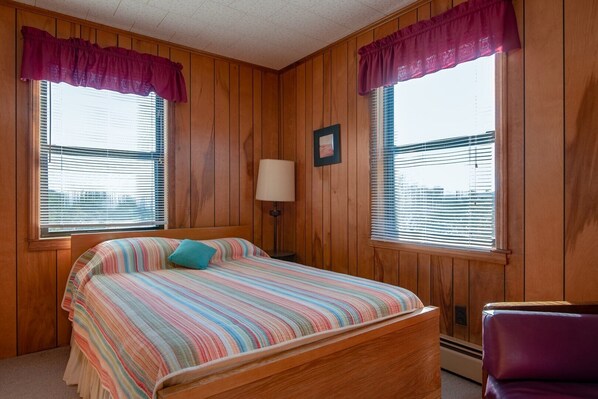Une chambre avec des lambris en bois, un lit recouvert d’une courtepointe à rayures, deux fenêtres équipées de stores, une lampe de chevet et une chaise.