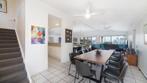 A spacious dining area with a long table and multiple chairs, a kitchen in the background, and a staircase leading to an upper level.