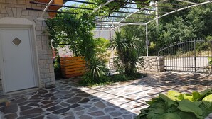 Een terras met stenen bestrating, een witte deur, een houten bank en weelderige beplanting.