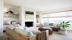 A cozy living room with a stone fireplace, a large window with a view of the ocean, and a comfortable seating area with a sofa and ottoman.