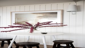 A dining area with a round white table, two dark stools, a vase with pink flowers, and a framed abstract painting on the wall.