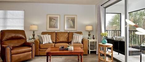 Un salón con un sillón de cuero marrón, una mesa de centro de madera, un sofá con cojines, dos lámparas de pared y un ventanal con vistas a palmeras.