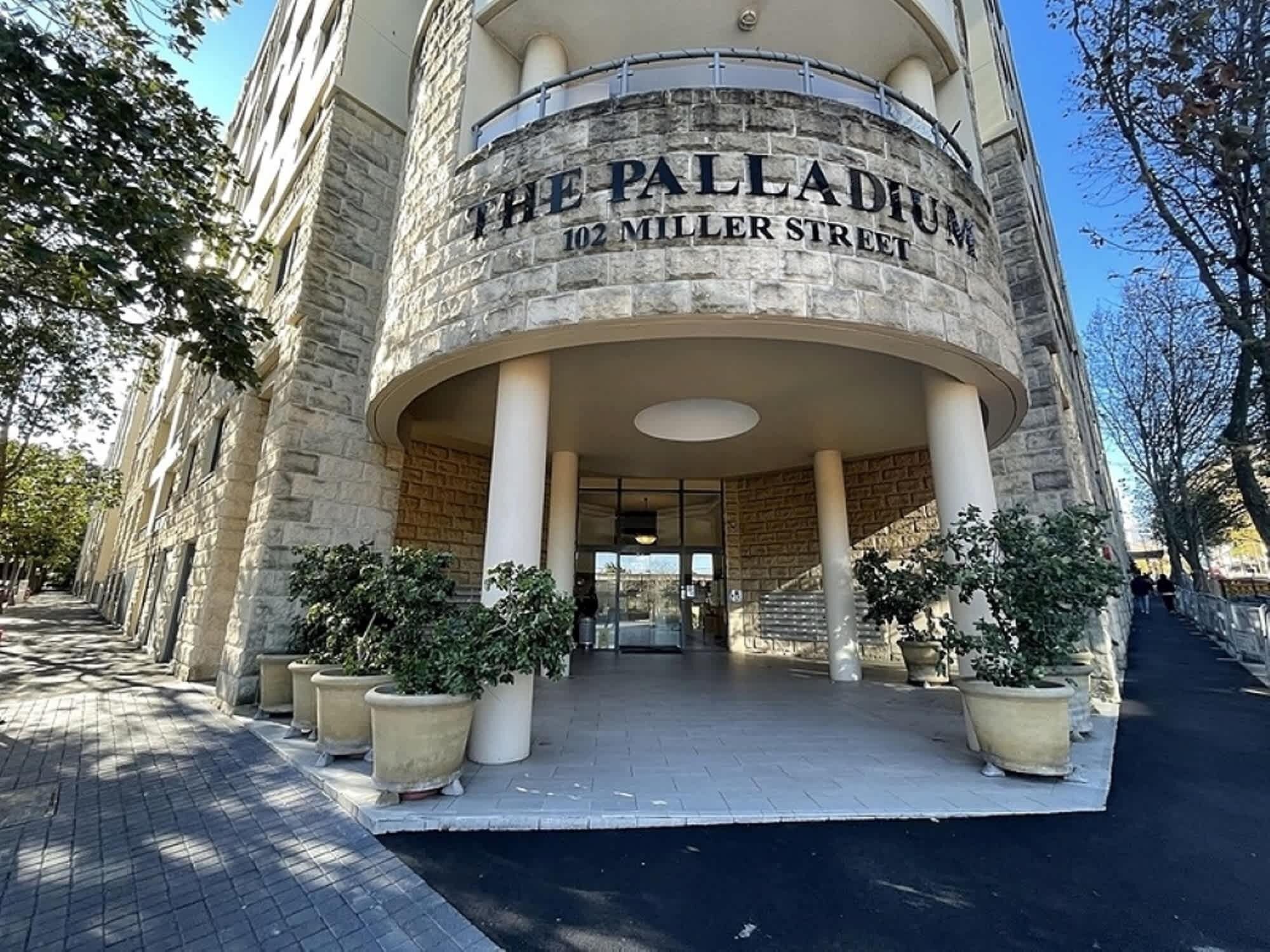 The Palladium building at 102 Miller Street with a stone facade, large columns, and potted plants at the entrance.