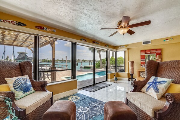 A spacious room with wicker furniture, large windows, a ceiling fan, and a view of a pool and palm trees.