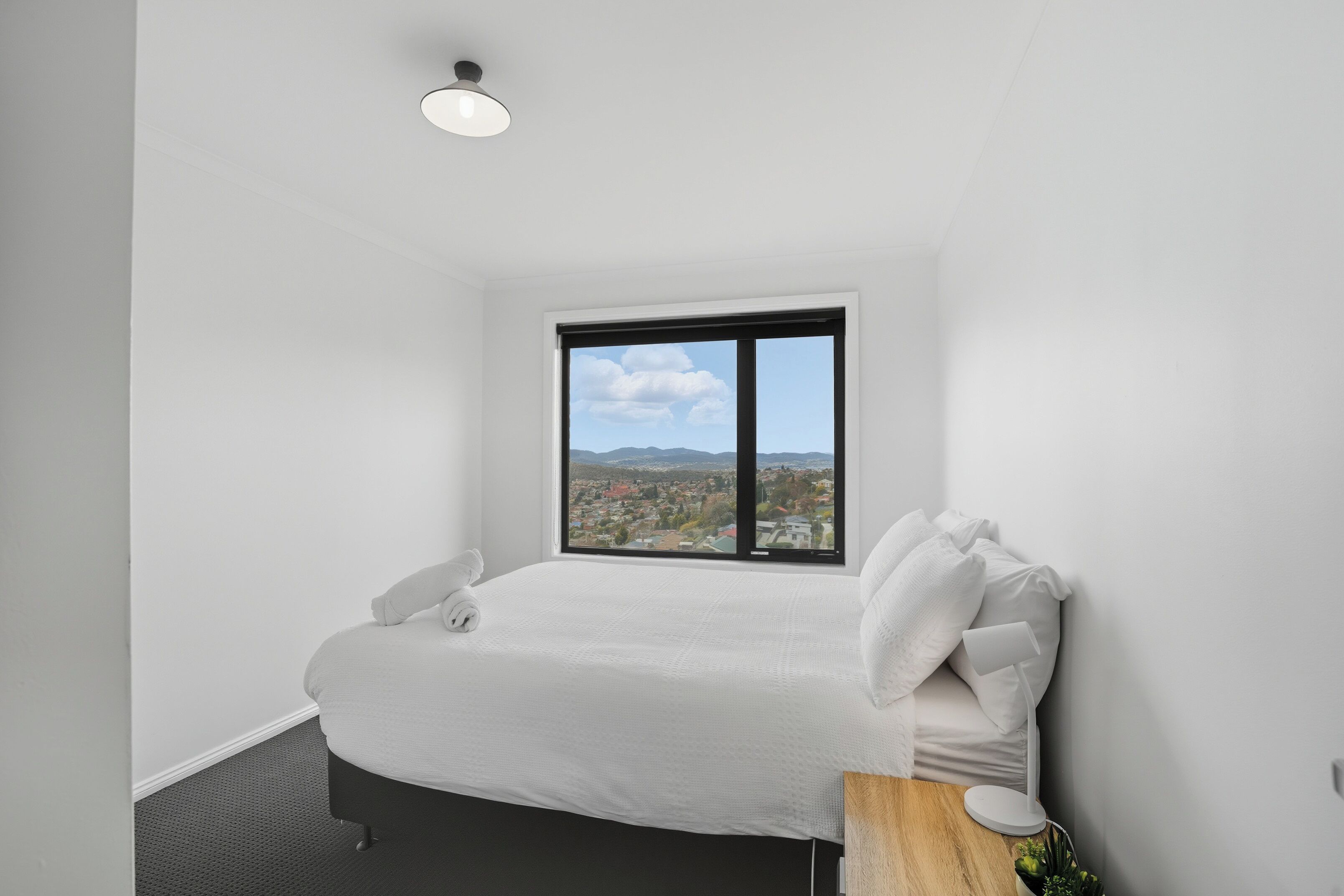 A bedroom with a bed, a window with a city view, a bedside table, and a lamp.