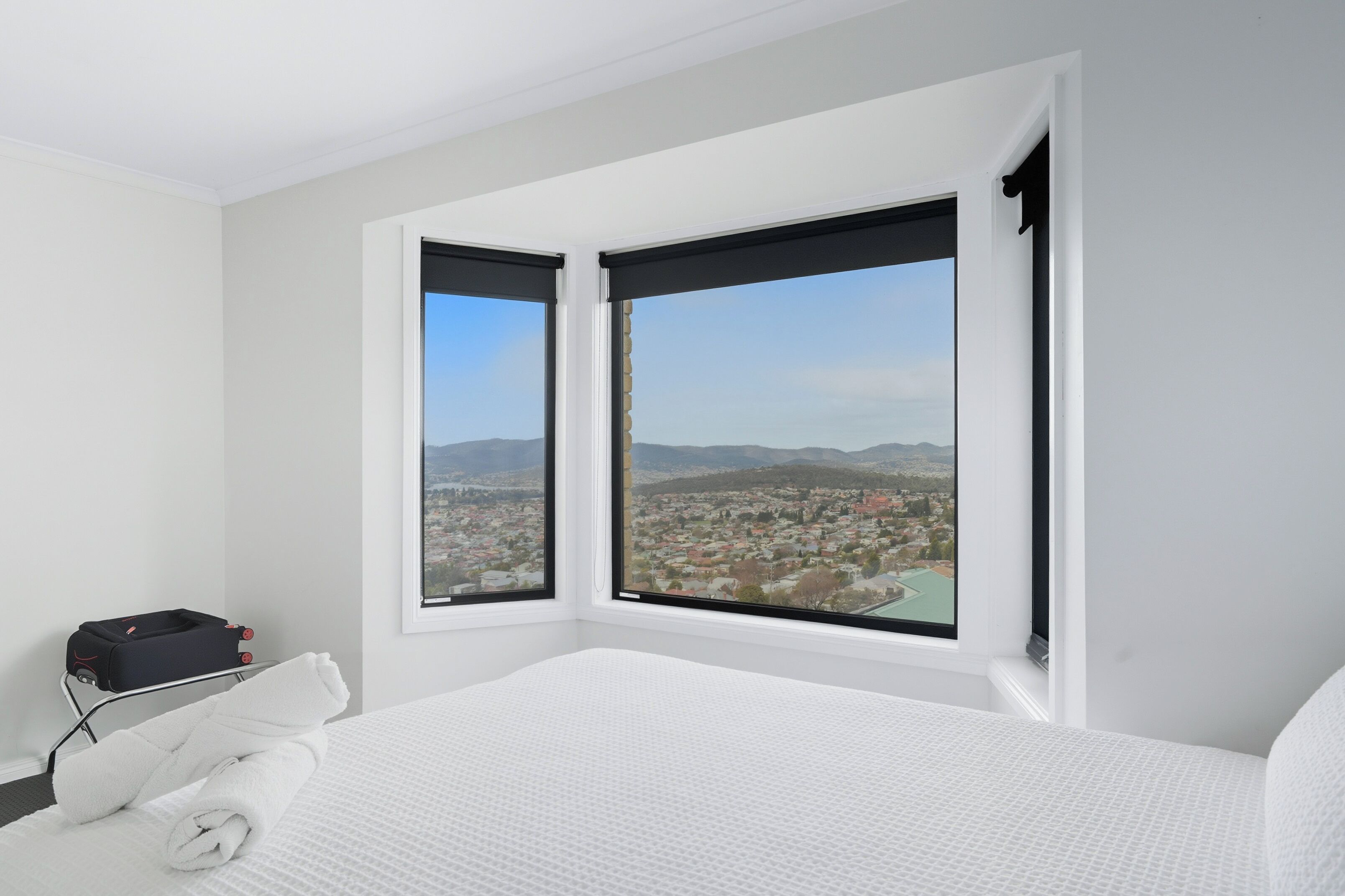 A hotel room with a large bed, two windows with black blinds, a view of a cityscape, and a small table with a suitcase and a towel.