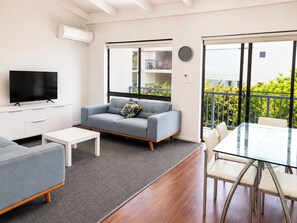 A modern living room with a flat-screen TV, grey sofa, white coffee table, and a glass dining table with chairs.