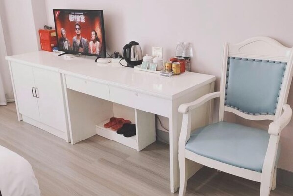 Un escriptori blanc amb una televisió, una papereta i diferents objectes. Hi ha una cadira blanca amb un coixí blau i una prestatgeria amb sabates sota l'escriptori.