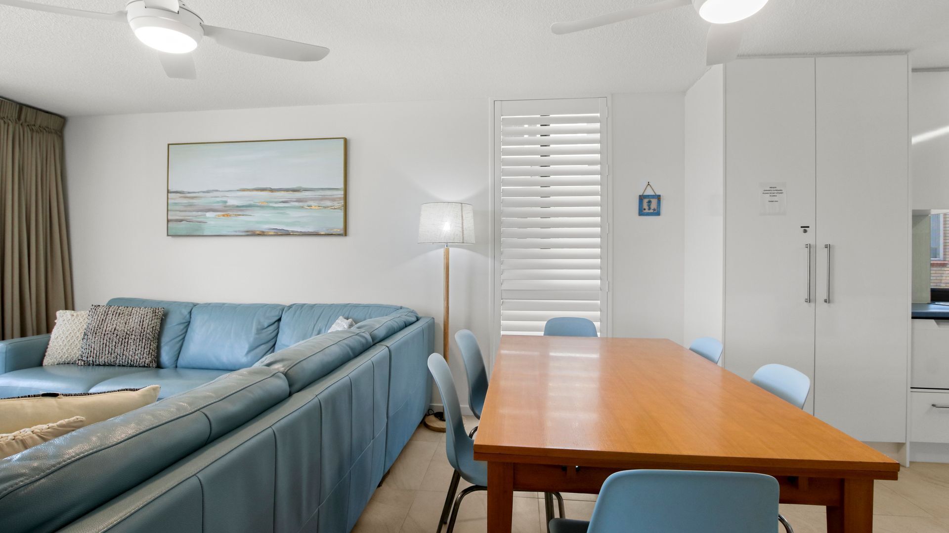 Un salon moderne avec un canapé bleu, une table à manger en bois et une grande fenêtre équipée de stores.