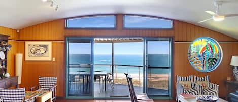 A spacious room with a large sliding glass door leading to a balcony with a view of the ocean. There is a dining area with checkered chairs and a table, and a colorful artwork on the wall.