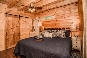 Une chambre dans une cabane en bois, comprenant un lit, une table de chevet, une commode et un ventilateur de plafond.
