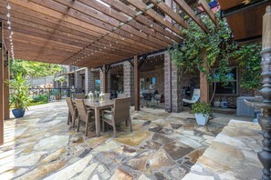 An outdoor patio with stone flooring, wooden tables, and chairs, surrounded by brick walls and greenery.