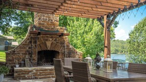 An outdoor stone fireplace with a pergola overhead, surrounded by wicker furniture and lanterns. - Your PRIVATE Hot Springs Lake Hamilton Vacation Paradise  (Royal)