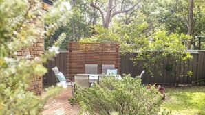 Una terraza de madera con una mesa y sillas, rodeada de follaje y un muro de ladrillo.