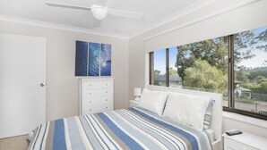 Un dormitorio con cama, cómoda, mesita de noche, ventilador de techo y ventana con vista a árboles y casas.