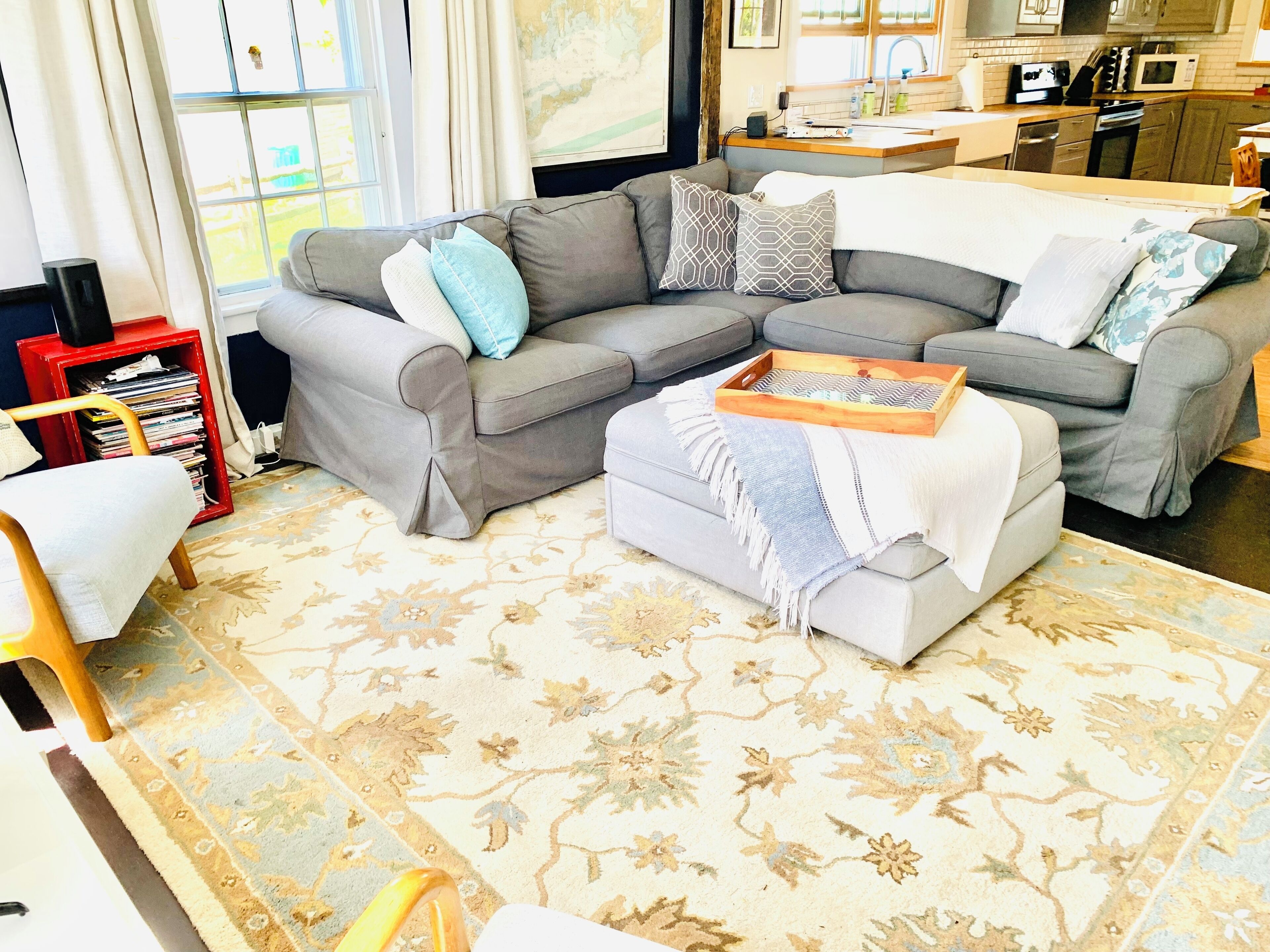 Ein Wohnzimmer mit einem grauen Ecksofa, einem kleinen Hocker und einem Holztablett. Es gibt ein rotes Bücherregal mit Zeitschriften sowie einen kleinen Tisch mit einer Lampe. Das Zimmer verfügt über ein großes Fenster mit Vorhängen und einen Blick ins Freie.