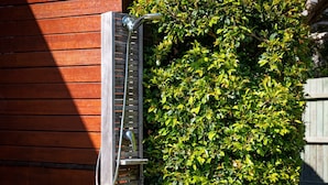 Une douche extérieure en métal, dotée d’un tuyau, est placée contre un mur en bois et entourée d’un feuillage verdoyant et luxuriant.