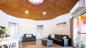 Un vaste espace de vie doté d’un plafond en bois, d’un canapé en cuir noir, d’une table à manger avec des chaises blanches et d’une porte en verre donnant sur l’extérieur.