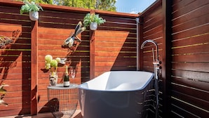 Une salle de bain extérieure moderne avec une baignoire sur pieds, une petite table avec une bouteille de champagne et un verre, et deux plantes en pot.