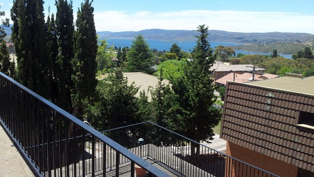 Un balcon doté d’une rambarde en métal, donnant sur un quartier résidentiel avec des maisons, des arbres et une étendue d’eau visible au loin.