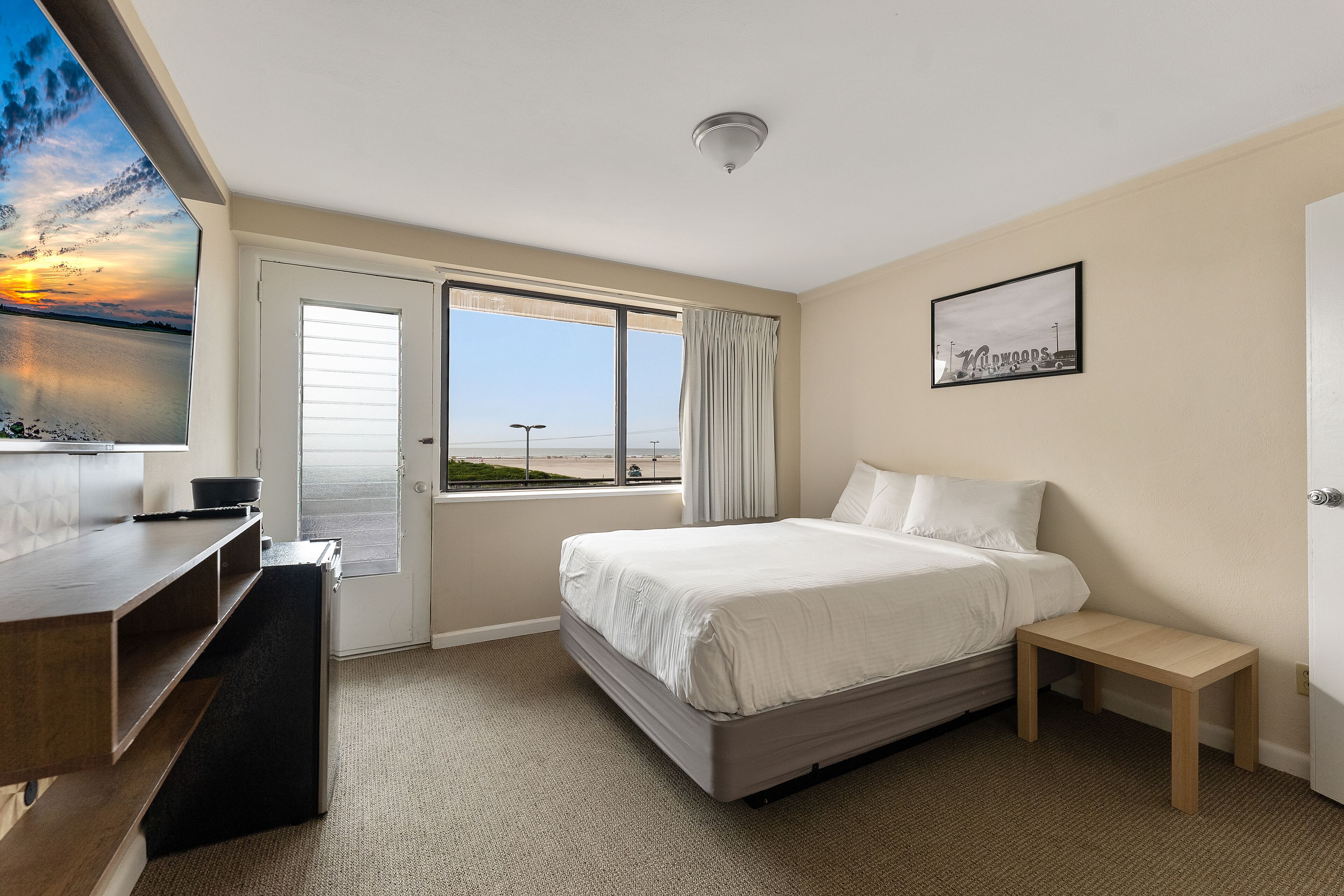 A hotel room with a bed, a desk, a small table, a window with curtains, and a painting on the wall.