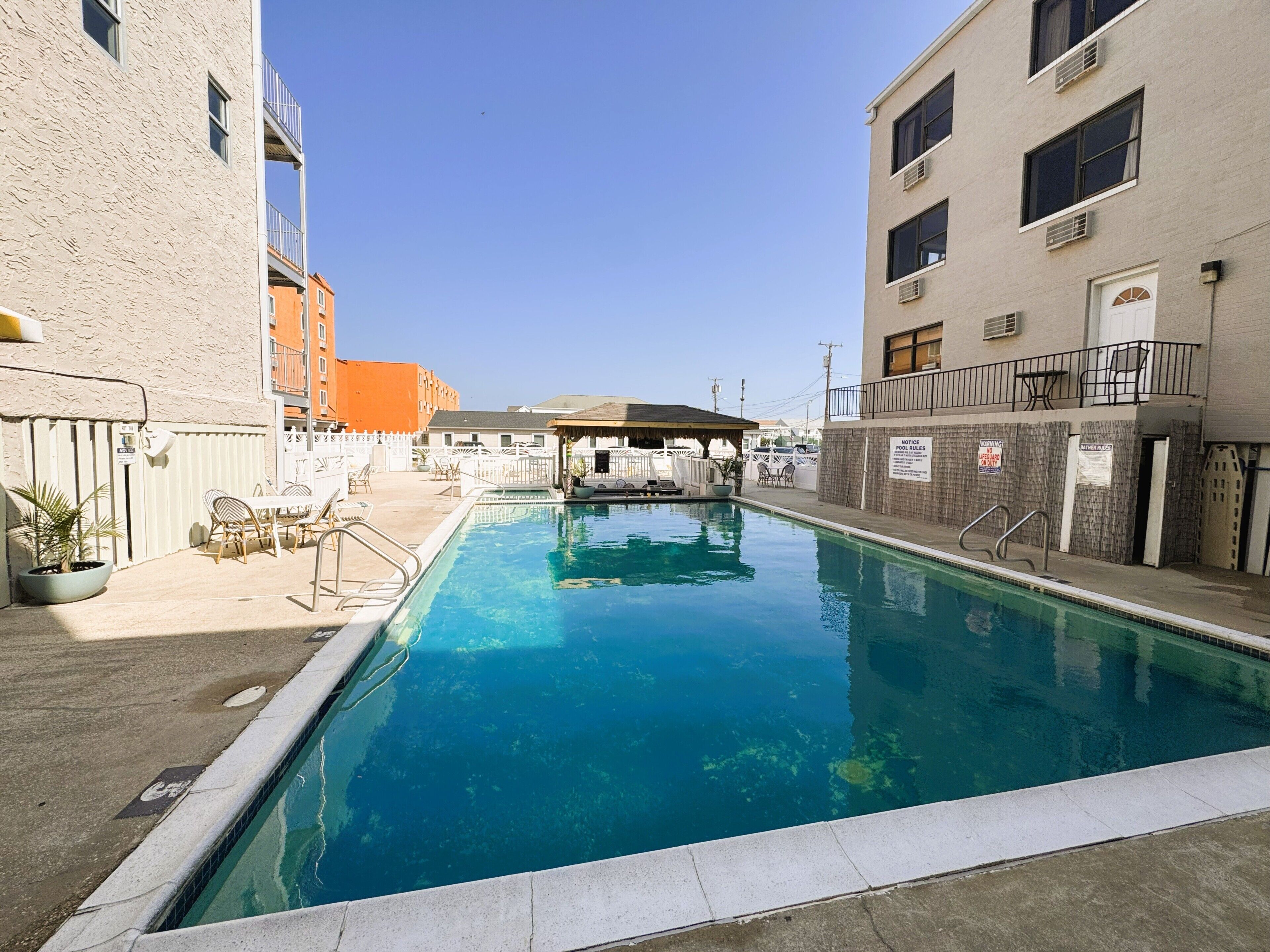 Uma piscina com espreguiçadeiras e mesas, cercada por edifícios com varandas e um céu claro.