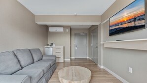 A modern living room with a grey sofa, a round wooden coffee table, and a flat-screen TV mounted on the wall displaying a sunset scene.
