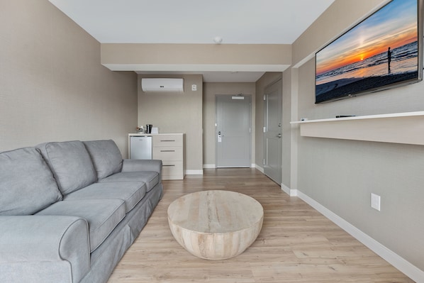 A modern living room with a grey sofa, a round wooden coffee table, and a flat-screen TV mounted on the wall displaying a sunset scene.
