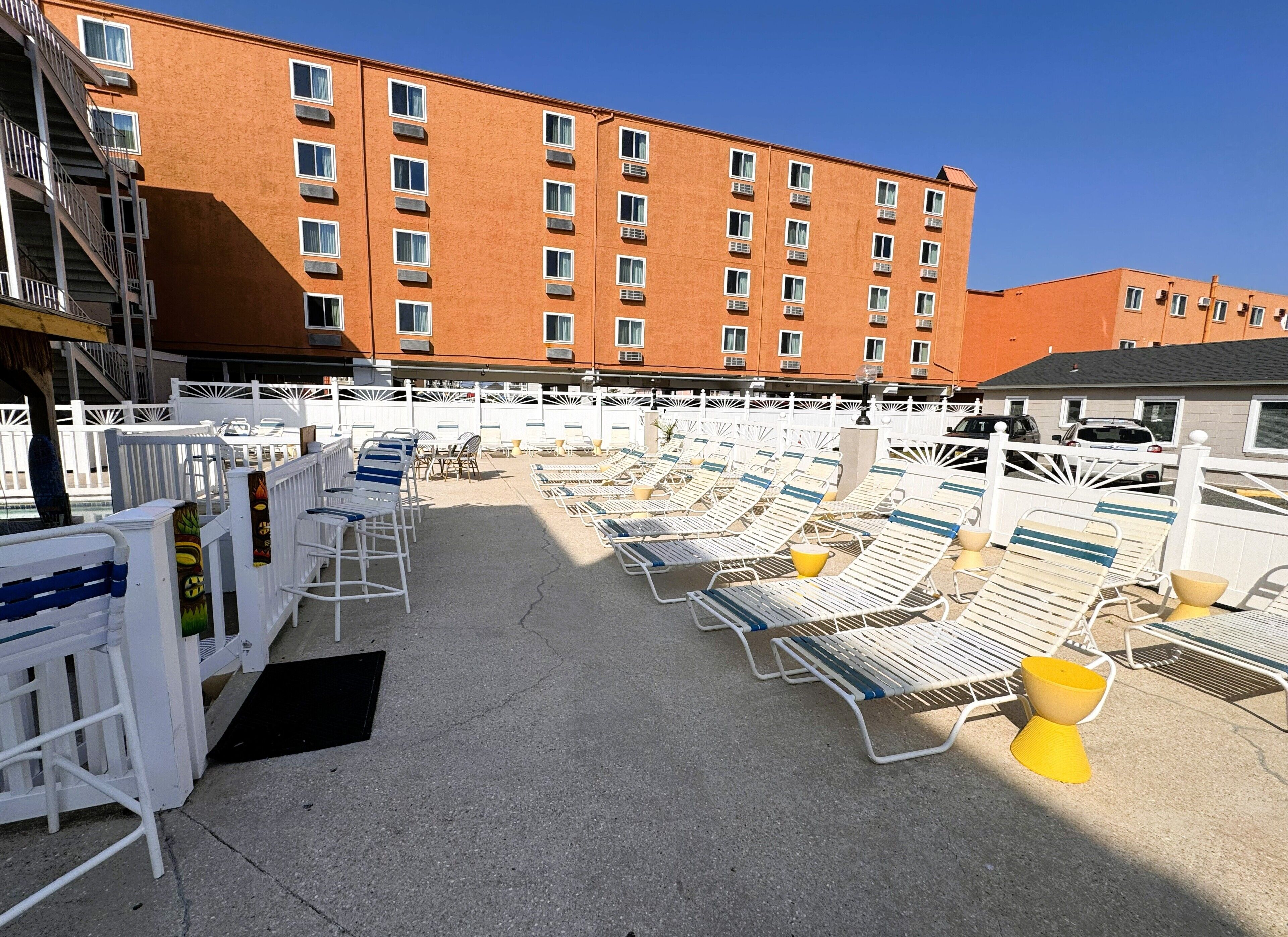 A rooftop pool area with sun loungers and a white fence, adjacent to a multi-story building with balconies.